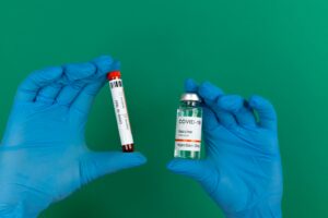 Hands in gloves holding COVID-19 vaccine and test vial on green background.
